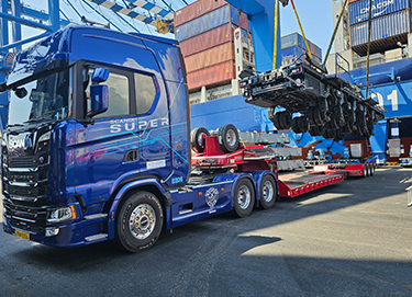 A truck at port is parked in the foreground. Stacks of shipping containers are in the background.