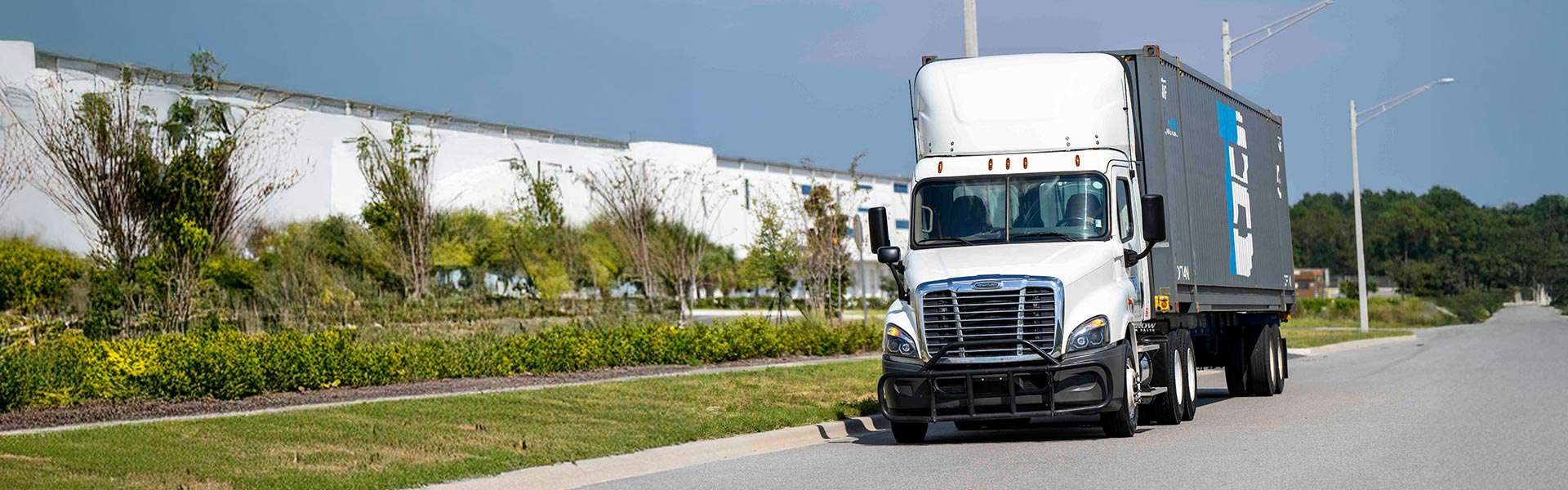 A Trailer Bridge truck drives along a quiet road, with a large white building in the background.