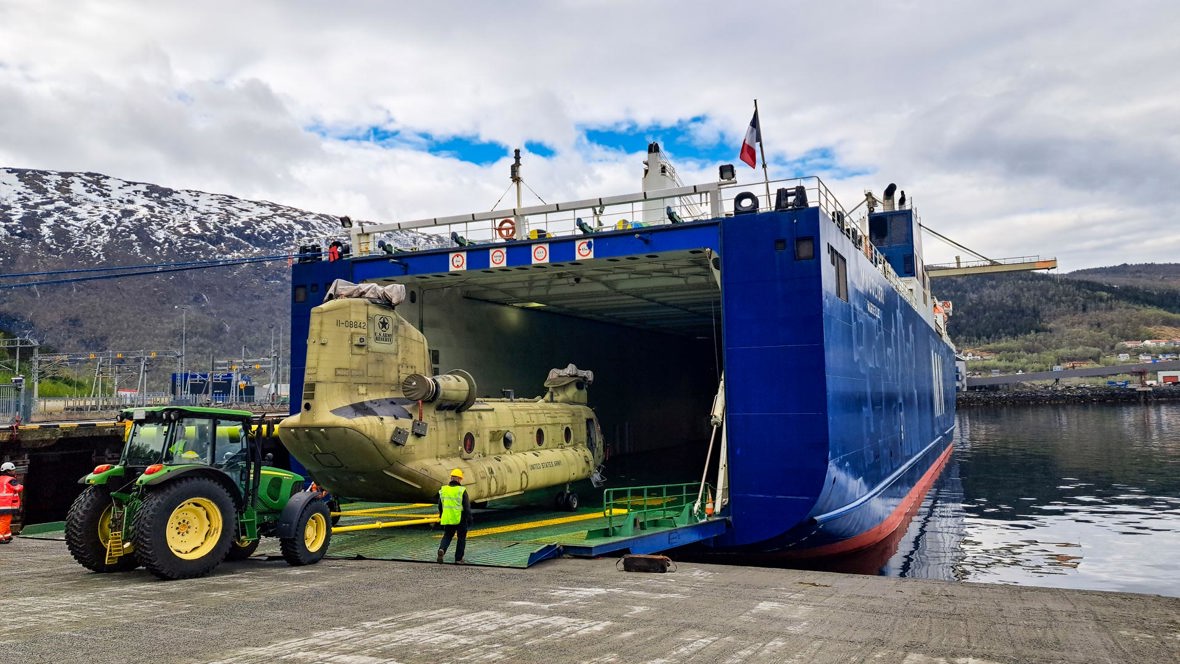 A military vehicle is loaded onto a ship, mountains in the background.
