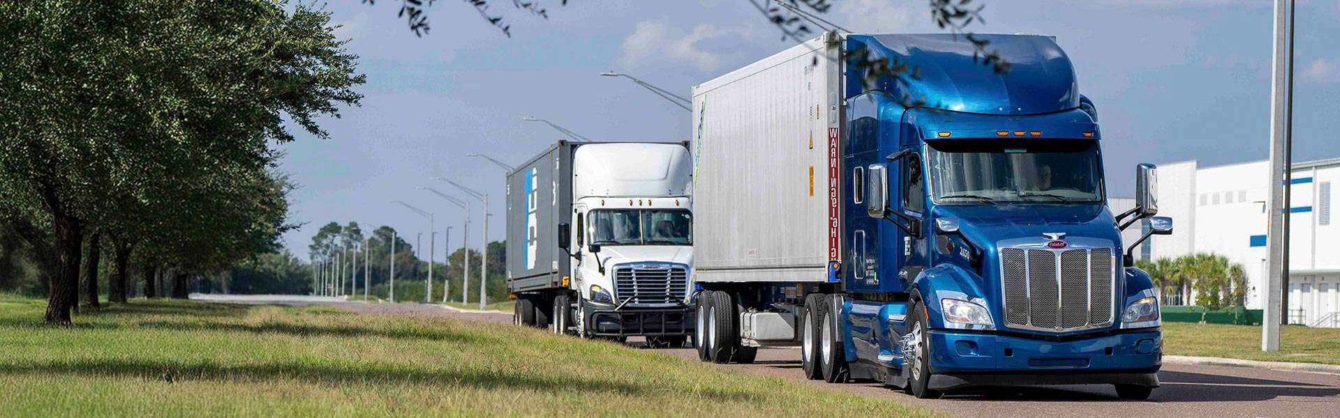 Two semi-trucks drive down a quiet street. The rear truck has the Trailer Bridge logo.