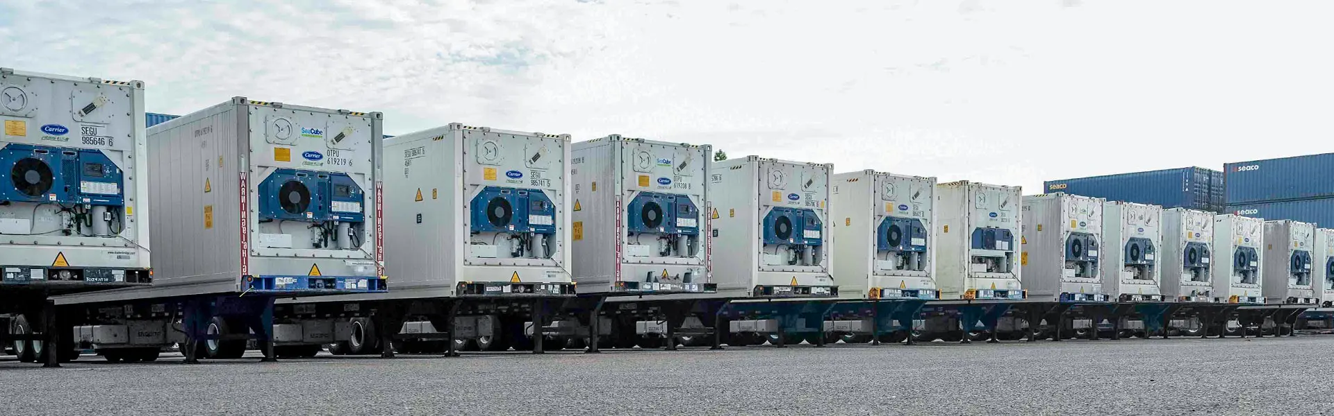 A line of refrigerated truck trailers are in the foreground and stacked containers are in the background.