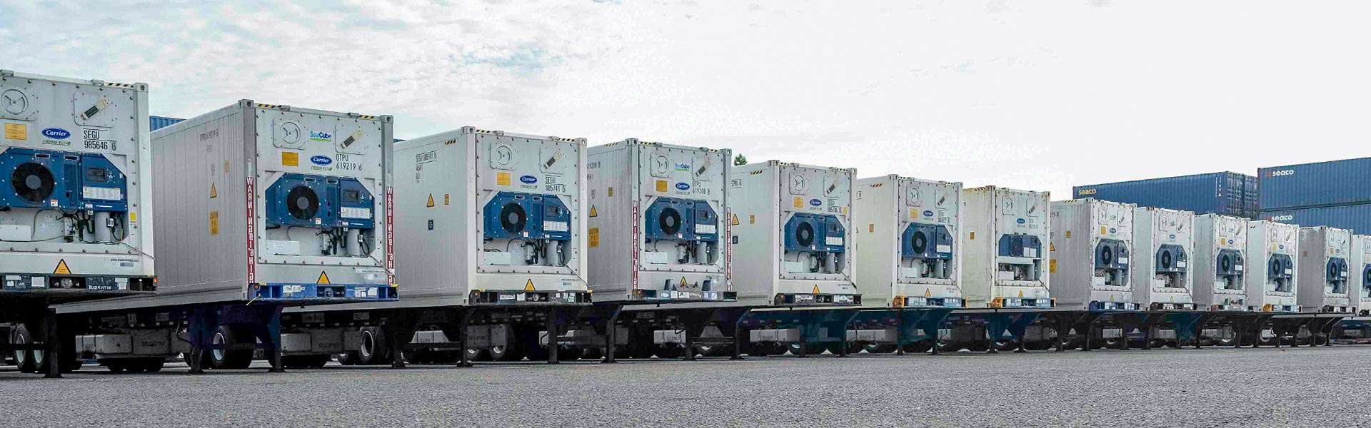 A line of refrigerated truck trailers are in the foreground and stacked containers are in the background.