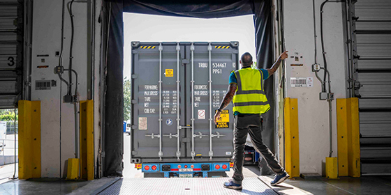 An employee with a high-visibility jacket directs a truck backing a trailer into a loading dock.
