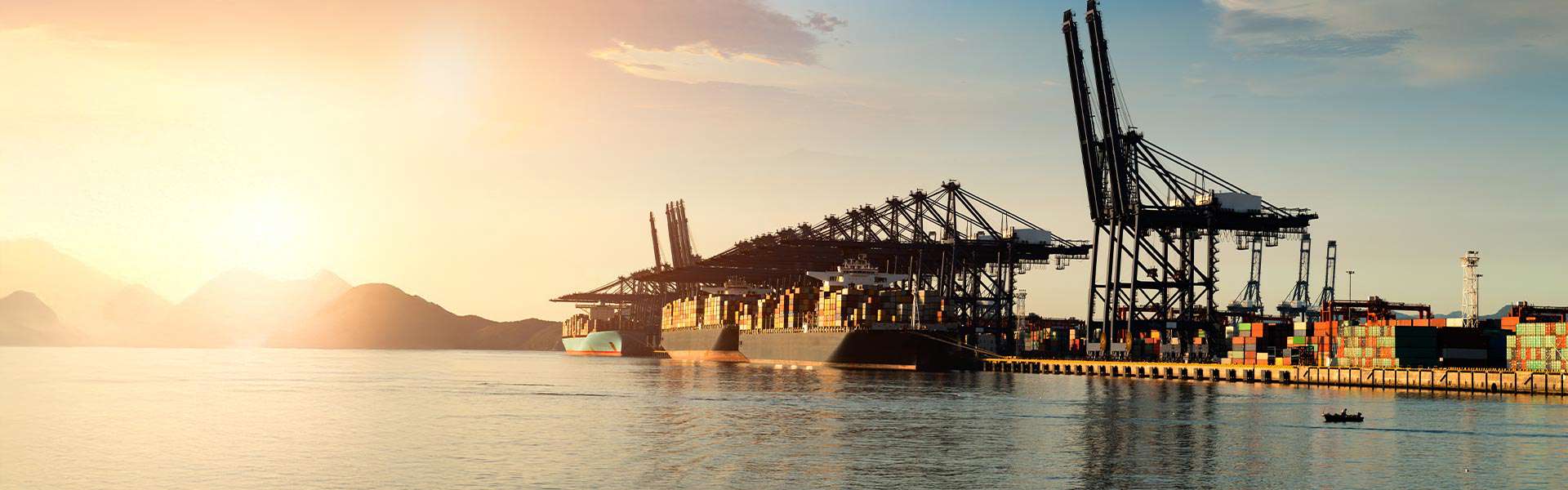 Barges are loaded with shipping containers in a port at sunrise or sunset.