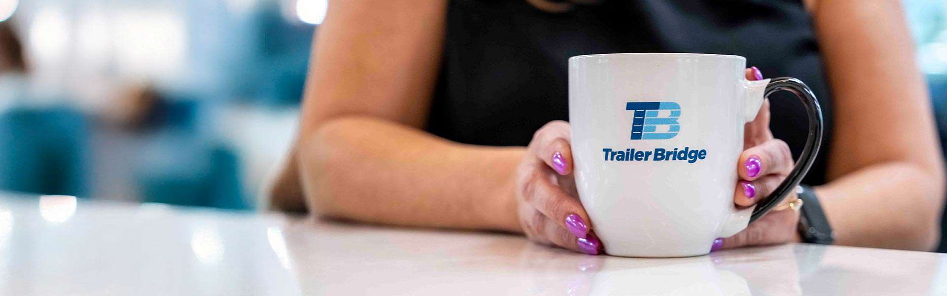 A person at a table holds a Trailer Bridge mug toward the camera.