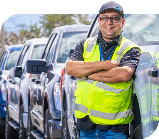 A Trailer Bridge employee wears a high visibility vest while leaning against a vehicle, with a line of other vehicles behind.