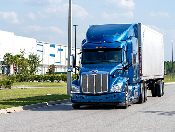 A truck drives down a quiet road, a warehouse building in the background.