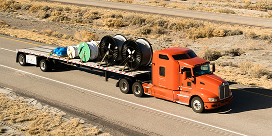 A flatbed truck drives along a desert-like road, carrying large spools of wire and cords.