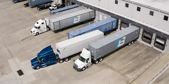 Semi-trucks leave loading docks with containers.