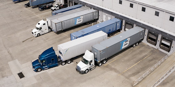 Semi-trucks leave loading docks with containers.