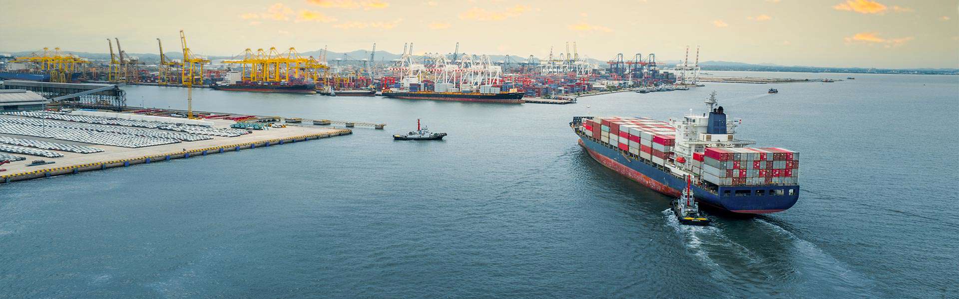 A barge full of shipping containers sails toward a busy port full of vehicles, cranes, and more containers.