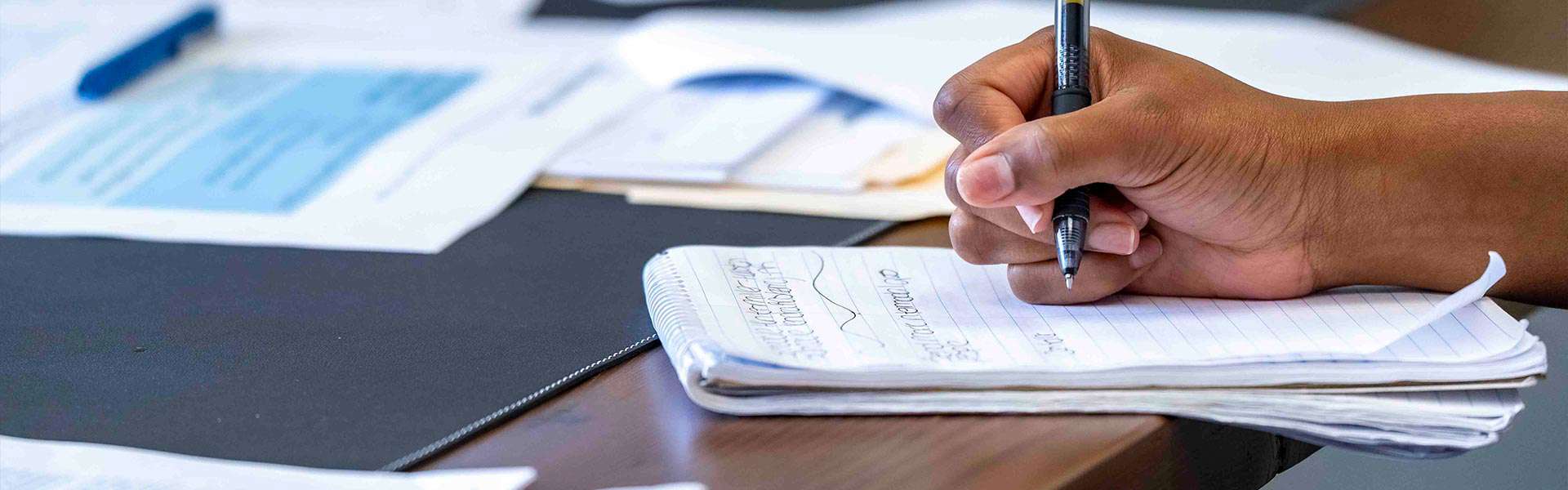 Close-up of a person writing in a notebook on a table covered with paperwork.