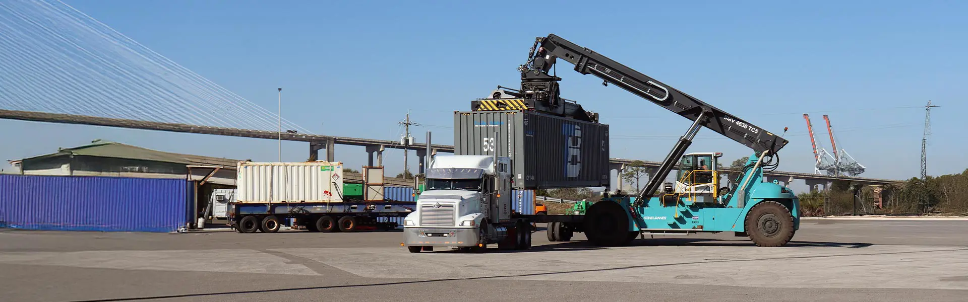 A crane lifts a container off a truck bed while more containers and trucks sit in the background.