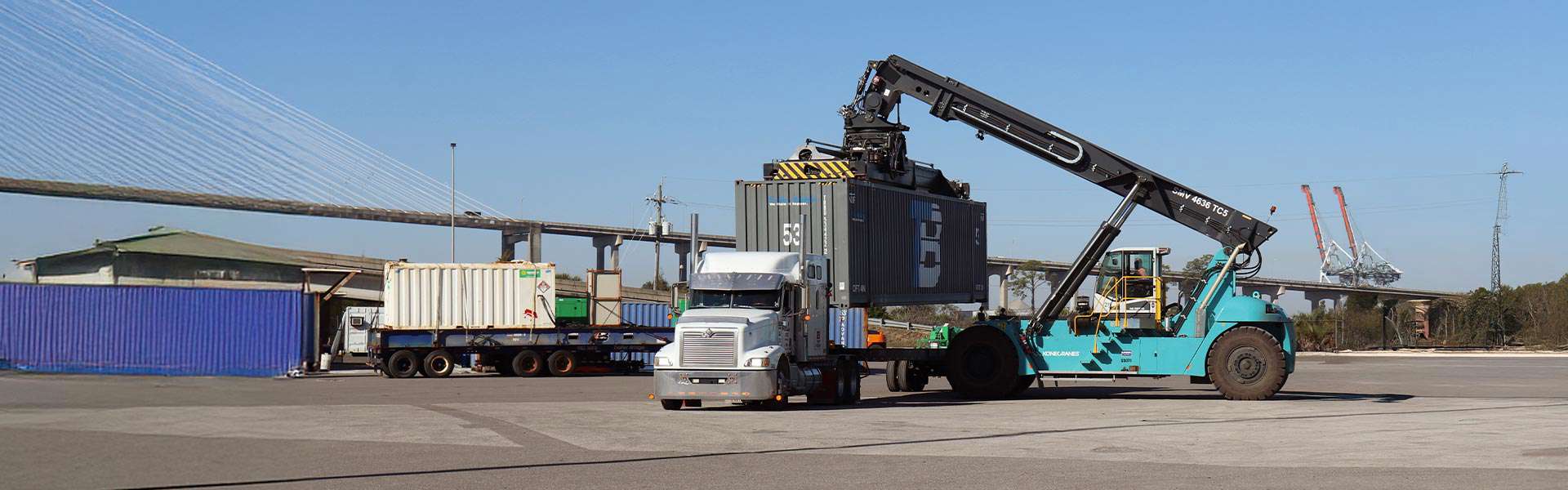A crane lifts a container off a truck bed while more containers and trucks sit in the background.