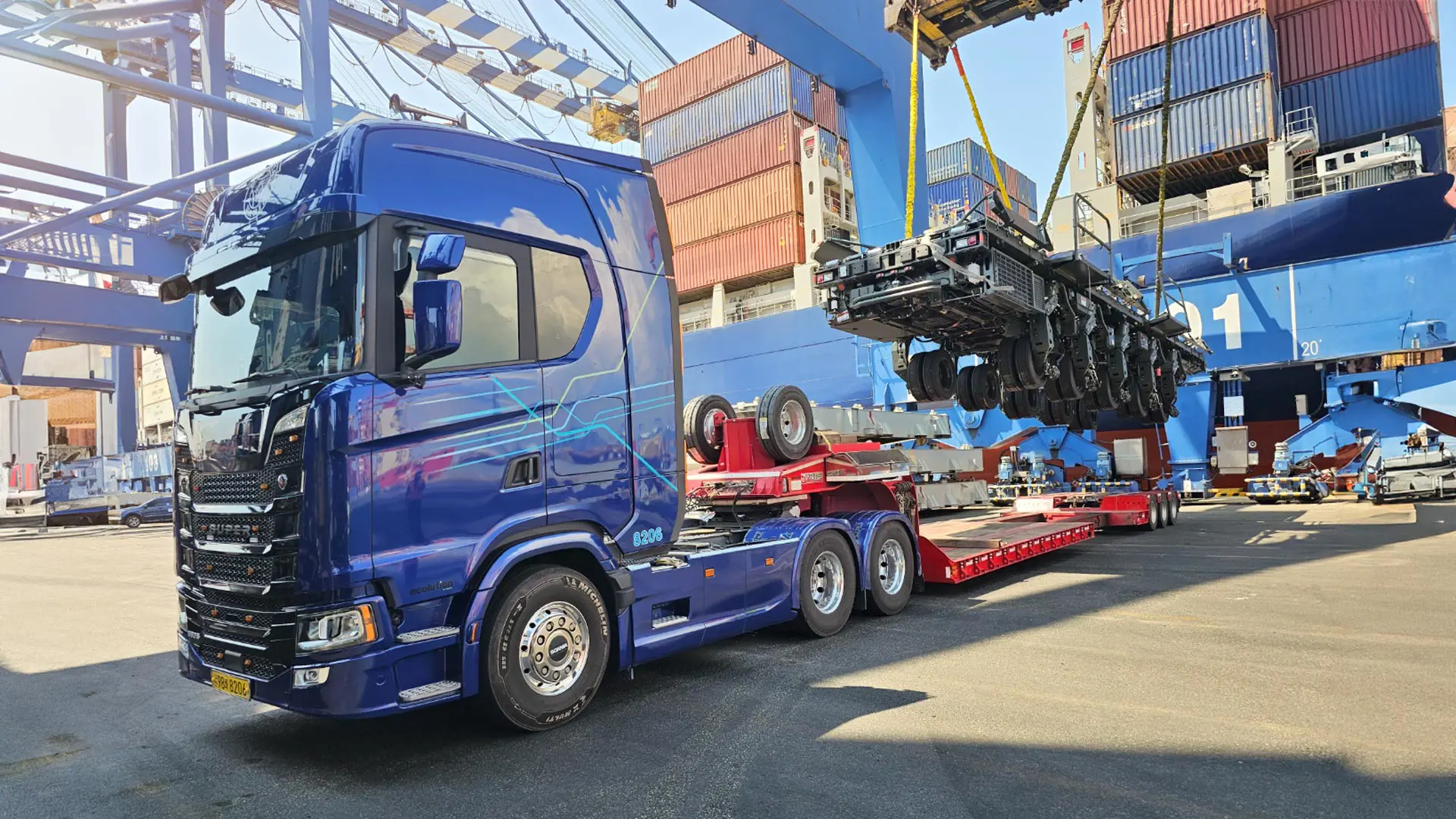 A truck at port is parked in the foreground. Stacks of shipping containers are in the background.