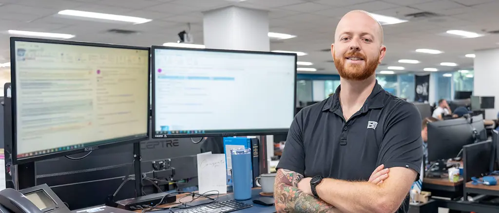 A Trailer Bridge employee smiles toward the camera and stands with arms crossed in front of two large computer monitors.
