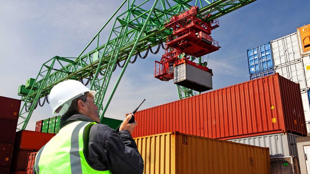 An employee in a high visibility vest instructs a crane moving shipping containers around a shipyard.
