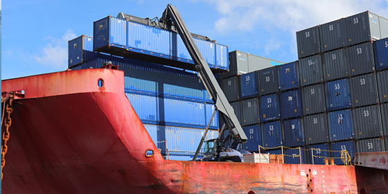 A crane moves a shipping container at the top of a stack of blue shipping containers.