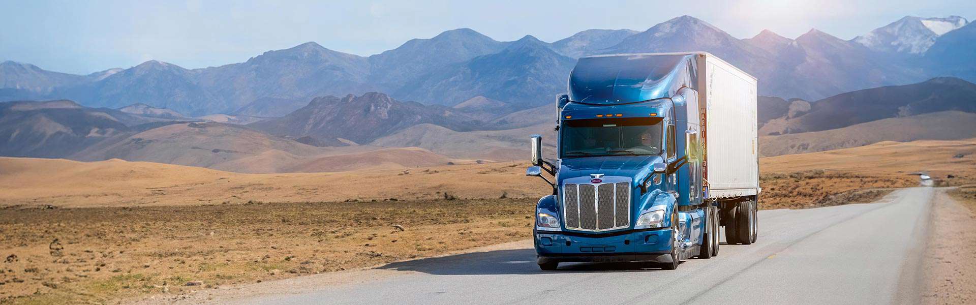 A semi-truck drives along a quiet road with mountains in the background.