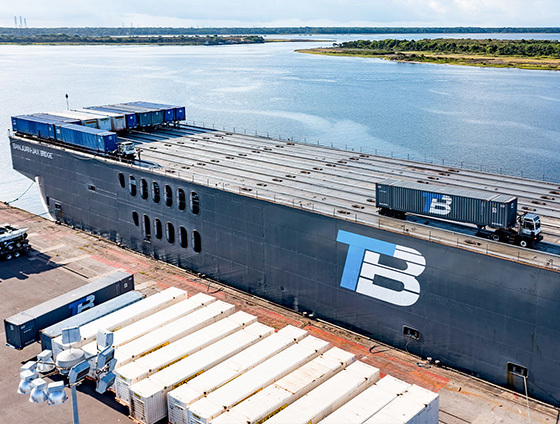 A barge with the Trailer Bridge logo is being loaded with shipping containers.
