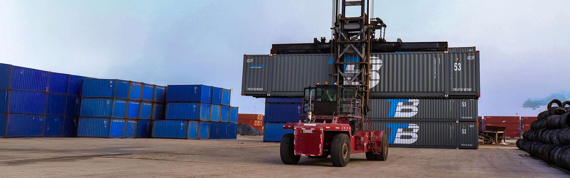 A crane moves the topmost shipping container on a stack of three shipping containers with the Trailer Bridge logo.