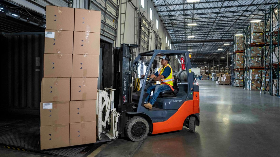 A forklift operator moves a stack of boxes in a warehouse.