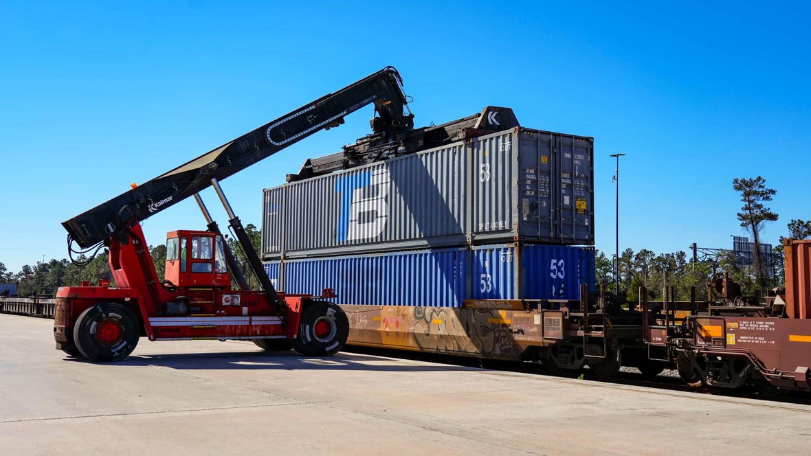 A crane vehicle lines up to move a shipping container off a train.