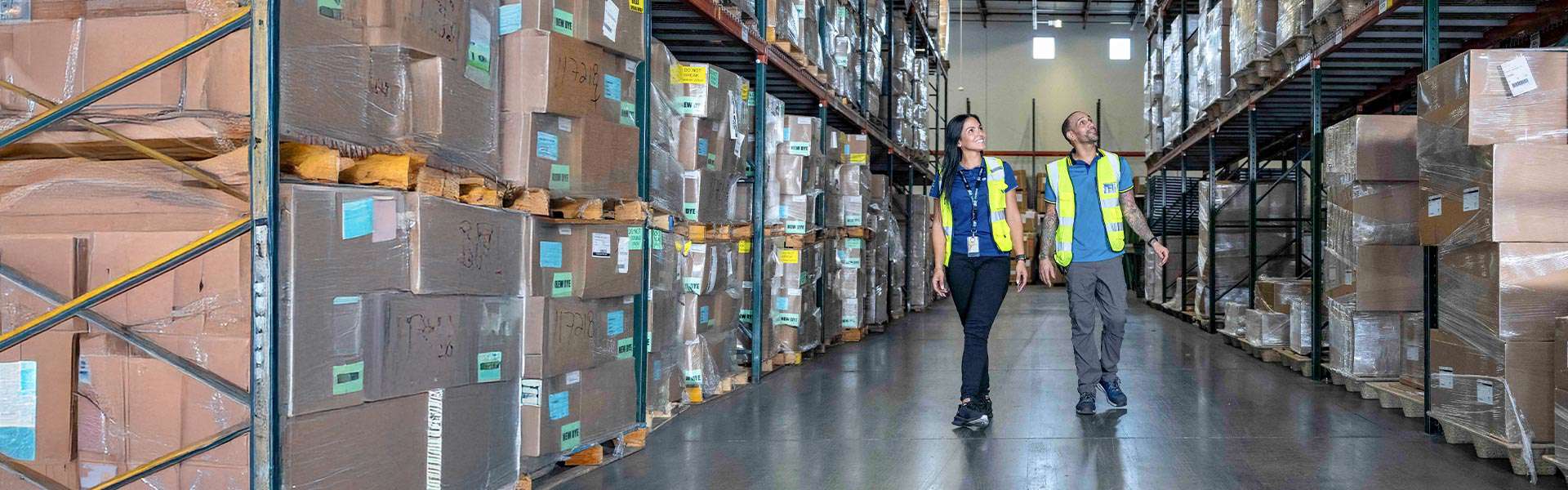 Two Trailer Bridge employees in high-visibility jackets walk around a warehouse stacked with cardboard boxes.