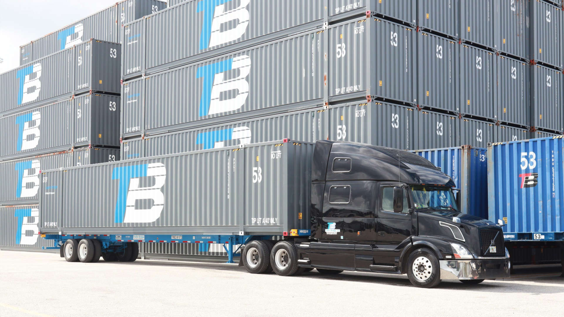 A semi-truck with a Trailer Bridge container is parked in front of stacks of other TB-branded containers.