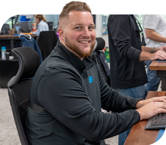 A Trailer Bridge employee smiles at the camera, hands on a keyboard, while others work on computers in the background.