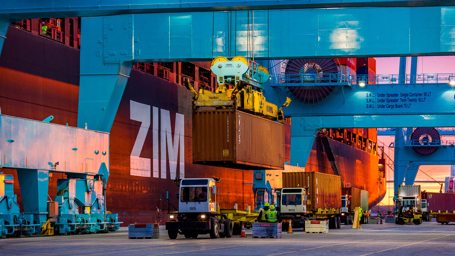 A crane unloads a shipping container from a truck while two others wait behind it for unloading.