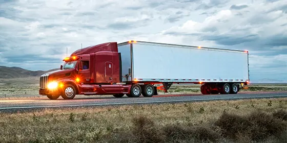 A semi-truck drives along a quiet road at twilight with lights on the front and the trailer.