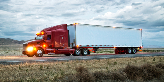 A semi-truck drives along a quiet road at twilight with lights on the front and the trailer.