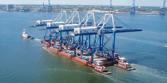 In the foreground is a container crane with a boat sailing away. A port is in the background.